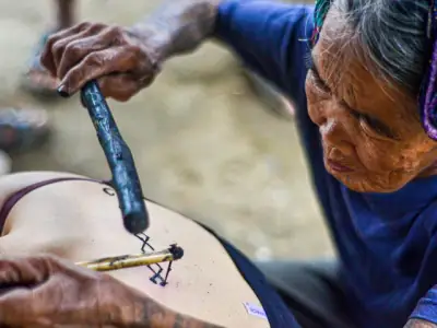 Elderly woman performing traditional hand-tap tattooing on a younger person's back in Kalinga, Philippines.