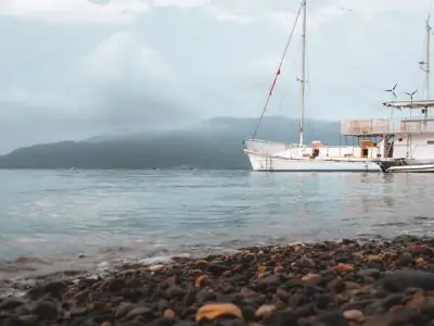 Tranquil view of a sailboat anchored near a stony beach in Batangas, Philippines under a misty sky.