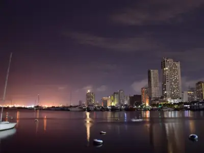 Low angle of modern boats moored in harbor of Manila with contemporary illuminated skyscrapers against evening sky